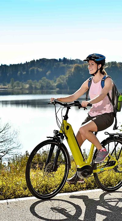 Incentives in Ostbayern - Radfahrerin mit E-Bike fährt auf Radweg bei sonnigem Wetter an einem See vorbei; Waldlandschaft im Hintergrund
